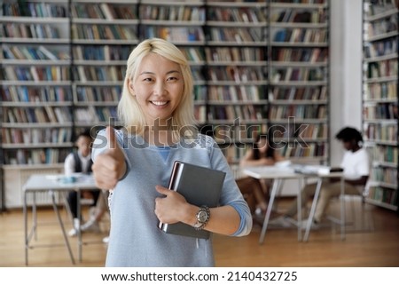 Asian student girl with blond dyed hair hold books pose in library showing thumb up at cam, mates on background. High school, university education, knowledge and studentship, excellent studies concept