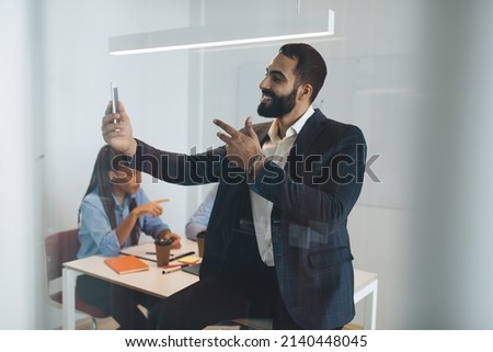 Cheerful Middle Eastern businessman in formal clothes taking selfie images on modern cellphone gadget using front camera, happy male employer making online video calling in office workspace