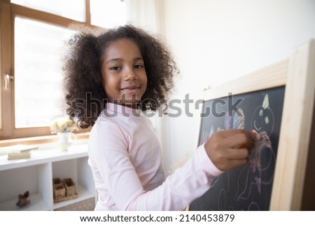 Happy American primary school kid drawing on blackboard, using colorful chalks, looking at camera, smiling. Kindergarten child hatching doodles on chalkboard with crayons. Head shot portrait