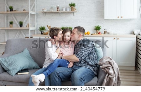 A family happily kisses a little girl at home, young parents and children enjoy loving hugs while sitting together on a sofa or sofa