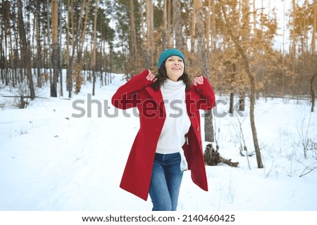 Cheerful beautiful mixed race woman in bright red coat spinning around herself feeling happy, enjoying freedom while relaxing in a snowy forest with sunbeams falling through pine trees Royalty-Free Stock Photo #2140460425