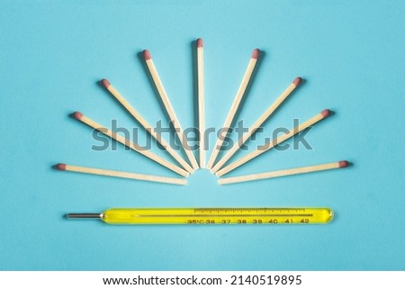 A group of matches ready to light forming a sun on top of a hot mercury thermometer on a colorful blue background. Conceptual photography, environment, and temperature.