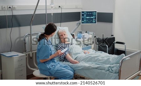 Nurse giving assistance to senior patient with disease in bed. Medical assistant and doctor doing healthcare checkup for pensioner with oxygen tube and IV drip bag in hospital ward. Royalty-Free Stock Photo #2140526701