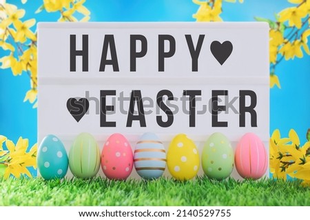 Bright positive spring Easter composition. Happy Easter text on lightbox against blue sky and yellow flowers. Multi-colored decorated eggs in a row on the grass.