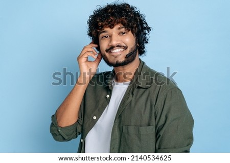 Portrait of friendly positive indian or arabian young adult man, call center, hotline employee with headphones and microphone, stands on isolated blue background, looks at the camera, smiling Royalty-Free Stock Photo #2140534625