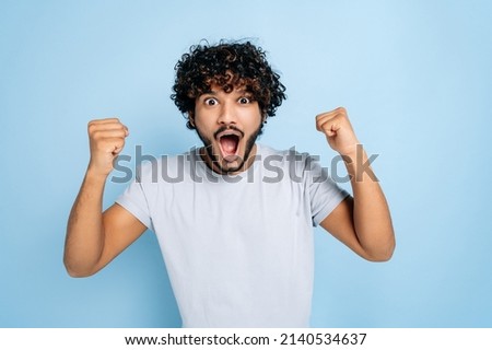 Enthusiastic amazed indian or arabic guy, in abasic t-shirt, rejoices in success, victory, win, gesturing with fists, looking at the camera, smiling, standing on an isolated blue background Royalty-Free Stock Photo #2140534637
