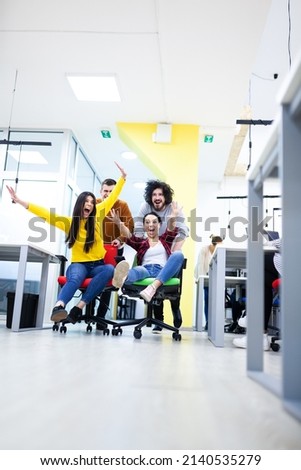 Colleagues having fun on work at the office. Girls pushing boys on chairs with wheels. Multiethnic group of successful start up company Royalty-Free Stock Photo #2140535279