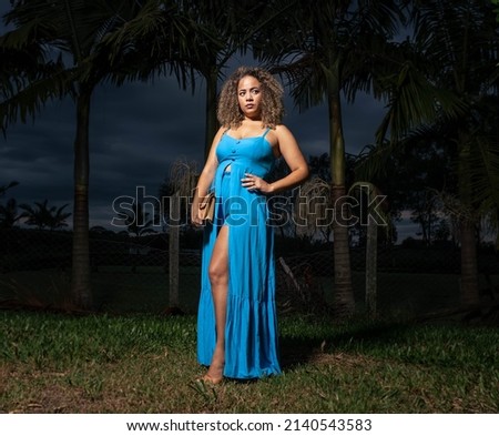 Brunette woman wearing a blue dress, beige bag and black boots on a green lawn area next to a lake looking towards the camera in an early evening with light from left to right.