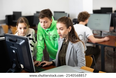 Friendly female teacher and positive teen students looking at monitor screen during lesson in computer lab at school library ..