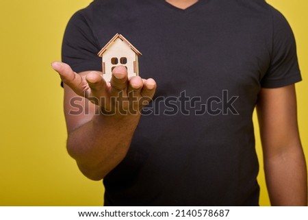 Photo of torso of black guy without head in frame in black everyday T-shirt, in his hand small toy house. Small house made of wooden construction set on palm of African-American on yellow background