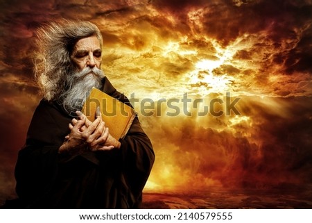 Prophet with Bible. Old Monk with Golden Book praying over Epic Landscape Background. Senior Bearded Man Worship in Black Cloak over Mystery Sunset Sky Royalty-Free Stock Photo #2140579555