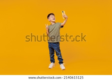 Full body little small happy boy 6-7 years old in green t-shirt do selfie shot on mobile cell phone show v-sign gesture isolated on plain yellow background. Mother's Day love family lifestyle concept