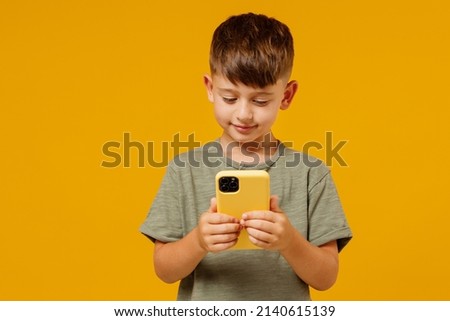 Little small smiling happy boy 6-7 years old wearing green t-shirt hold in hand use mobile cell phone isolated on plain yellow background studio portrait. Mother's Day love family lifestyle concept