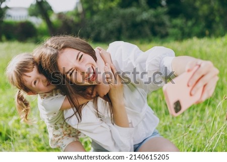 Smiling happy young woman in white clothes do selfie shot on mobile cell phone hug cuddle with child baby girl 5-6 years old Mommy rest with little kid daughter outdoor together. Love family concept.
