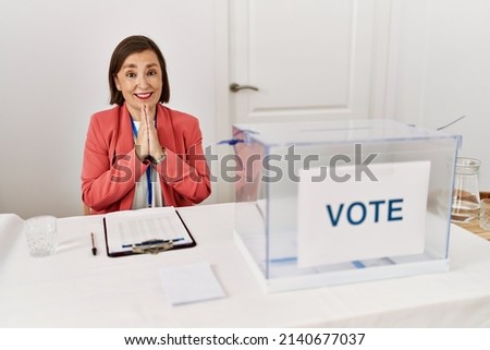 Beautiful middle age hispanic woman at political election sitting by ballot praying with hands together asking for forgiveness smiling confident. 