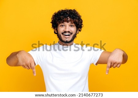 Amazed indian or arabian cheerful young man in white basic t-shirt looks at the camera and points fingers down at space for your presentation, stands on isolated orange color background, smiling Royalty-Free Stock Photo #2140677273