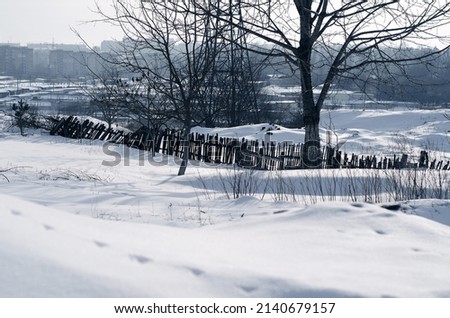 Winter landscape of outskirts of city. Leafless trees, snow, broken wooden fence. In distance city.