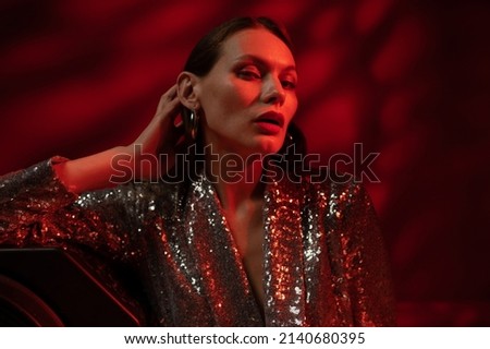 Fashion, beauty studio portrait of confident woman with wearing trendy sequin dress, silver hoop earrings, posing in red light. Copy, empty space for text