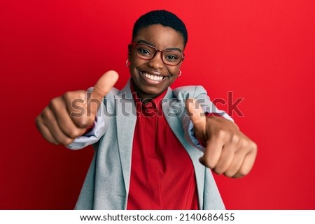 Young african american woman wearing business jacket and glasses approving doing positive gesture with hand, thumbs up smiling and happy for success. winner gesture.  Royalty-Free Stock Photo #2140686455
