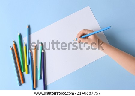 Child's hand with blank sheet of paper and different pencils on color background