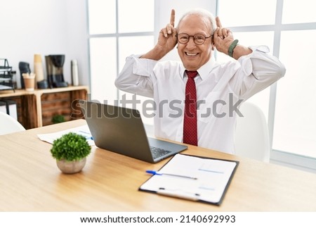 Senior man working at the office using computer laptop posing funny and crazy with fingers on head as bunny ears, smiling cheerful 
