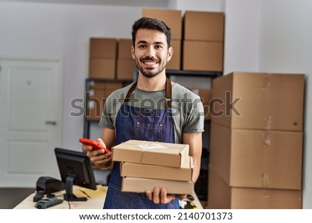 Young hispanic man business worker using smartphone holding packages at storehouse Royalty-Free Stock Photo #2140701373