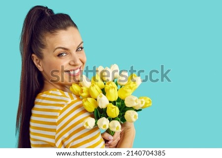 Studio portrait of a cheerful dark-haired young woman wearing a white shirt with yellow stripes stands half a turn with a bouquet of yellow tulips isolated on yellow background.