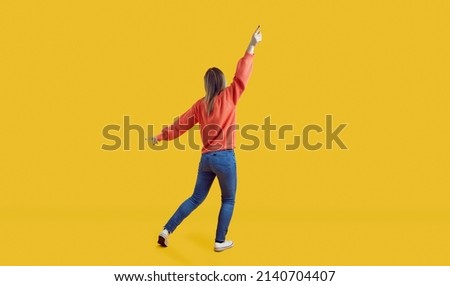 Happy woman having fun in studio. Cheerful excited carefree young girl in casual wear running and dancing isolated on vibrant yellow colour background. Full body shot, backside rear view from behind