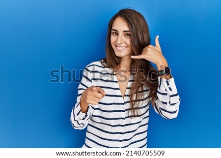 Young hispanic woman standing over blue isolated background smiling doing talking on the telephone gesture and pointing to you. call me. 