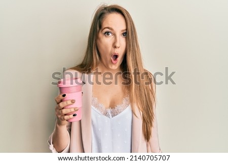 Young blonde woman drinking a take away cup of coffee scared and amazed with open mouth for surprise, disbelief face 