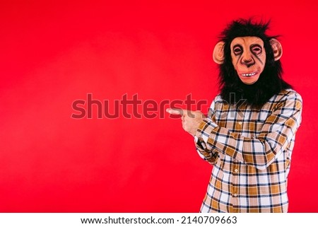 Man with chimpanzee monkey mask and plaid shirt, pointing fingers at copy space, on red background.