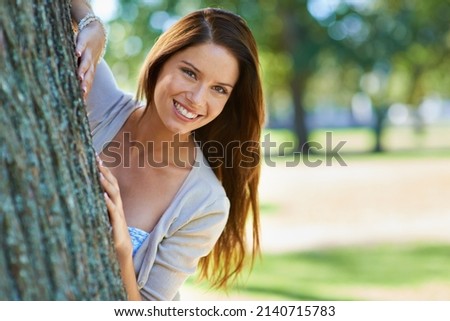 A nature lover. Shot of a beautiful young woman standing behind a tree at the park.