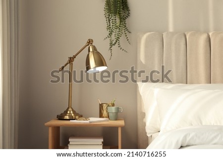 Stylish golden lamp and stationery on wooden nightstand in bedroom. Interior element Royalty-Free Stock Photo #2140716255
