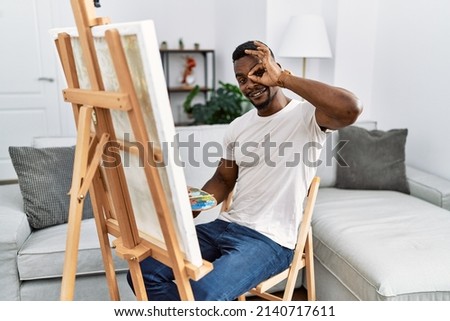 Young african man painting on canvas at home doing ok gesture with hand smiling, eye looking through fingers with happy face. 