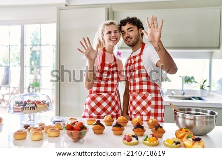 Couple of wife and husband cooking pastries at the kitchen showing and pointing up with fingers number ten while smiling confident and happy. 