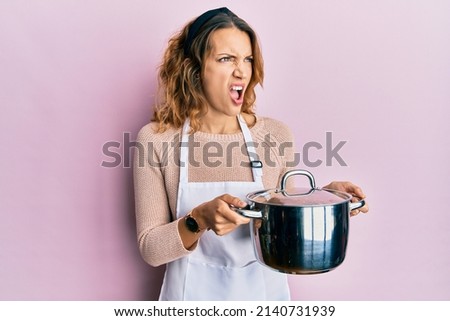 Young caucasian woman wearing apron holding cooking pot angry and mad screaming frustrated and furious, shouting with anger. rage and aggressive concept.  Royalty-Free Stock Photo #2140731939