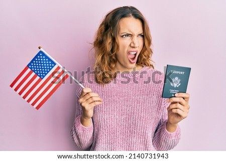 Young caucasian woman holding united states flag and passport angry and mad screaming frustrated and furious, shouting with anger. rage and aggressive concept.  Royalty-Free Stock Photo #2140731943