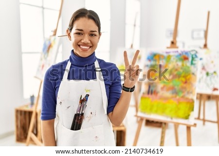 Young brunette woman at art studio showing and pointing up with fingers number two while smiling confident and happy. 