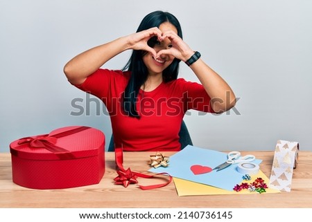 Beautiful hispanic woman with nose piercing doing handcraft creative decoration doing heart shape with hand and fingers smiling looking through sign 