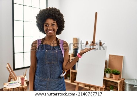 Young african american woman with afro hair at art studio smiling cheerful presenting and pointing with palm of hand looking at the camera. 