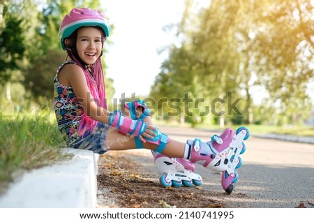 Laughing loudly, a funny girl with Afro-pigtails and wearing sports protective gloves and a helmet sat down to rest on the sidewalk after roller skating. A child's favorite hobby is roller skates. Royalty-Free Stock Photo #2140741995