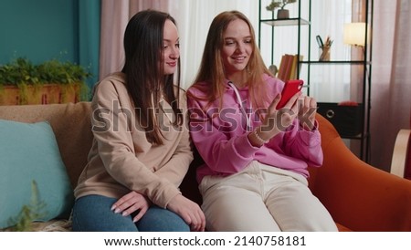 Cheerful girls friends siblings watching photos on smartphone, online dating app. Happy two female women couple family on sofa at home, enjoy chatting, spend leisure time on social media network