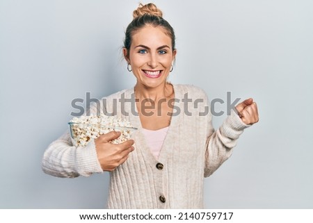 Young caucasian woman eating popcorn screaming proud, celebrating victory and success very excited with raised arm 