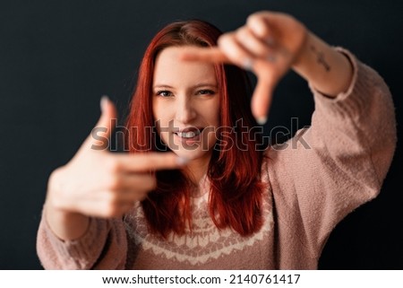 a beautiful and natural smiling red-haired skinny girl in a knitted sweater makes an imaginary lens or camera frame gesture with her fingers and looks through it at the viewer