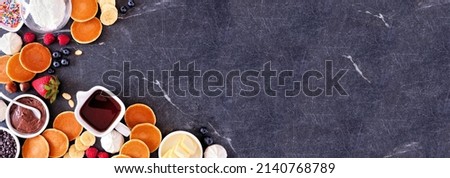 Breakfast or brunch pancake buffet corner border. Top view on a dark stone banner background. Mini pancakes with a selection of toppings.