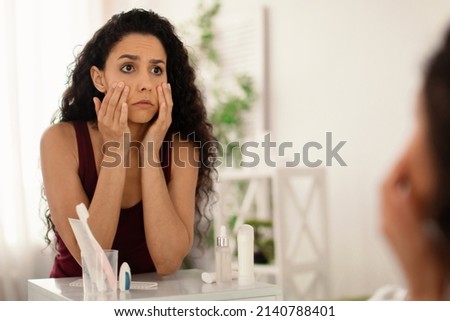 Beauty and skincare. Upset young woman looking in mirror and touching face, examining wrinkles and dark circles under her eyes at home, copy space. Dull tired skin concept