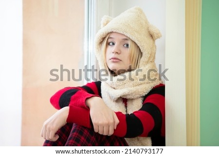 A teenage girl in a funny fur hat sits on the windowsill and looks at the camera.