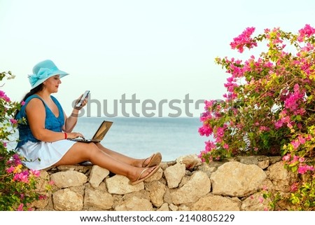 A young brunette woman in a hat sits on the background of the sea drinks a cocktail and works remotely on a laptop. Horizontal photo