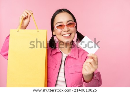 Happy young asian woman showing credit card for shopping, holding bag, buying on sale, going to the shop, store, standing over pink background