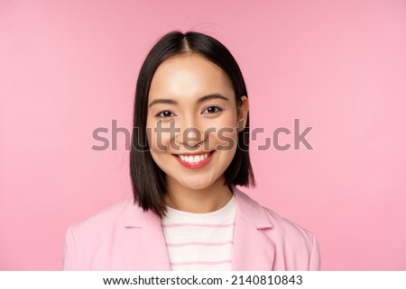 Close up portrait of asian corporate woman, looking professional, smiling at camera, wearing suit, standing over pink background